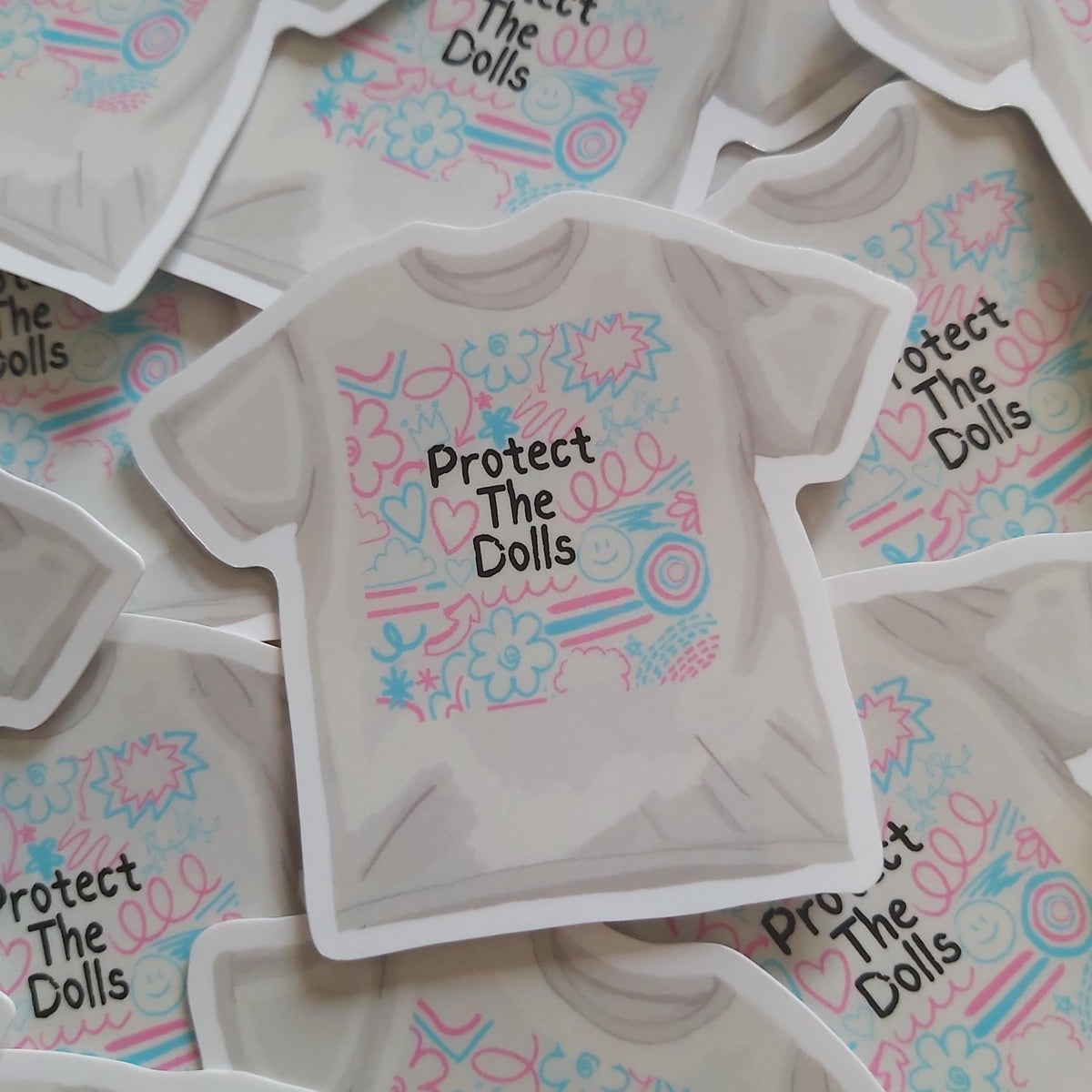 A die-cut sticker shaped like a short-sleeve T-shirt, featuring the text 'Protect The Dolls' in black alongside pink and blue retro doodles, displayed on a pile of similar stickers.