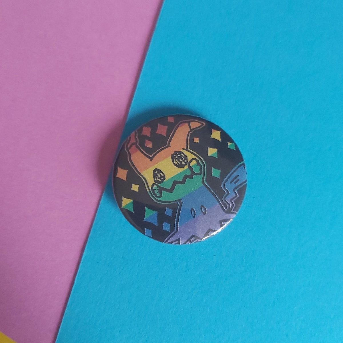 A round pin featuring a stylised Pokémon character with a rainbow background, resting on a diagonal split of pink and bright blue paper.