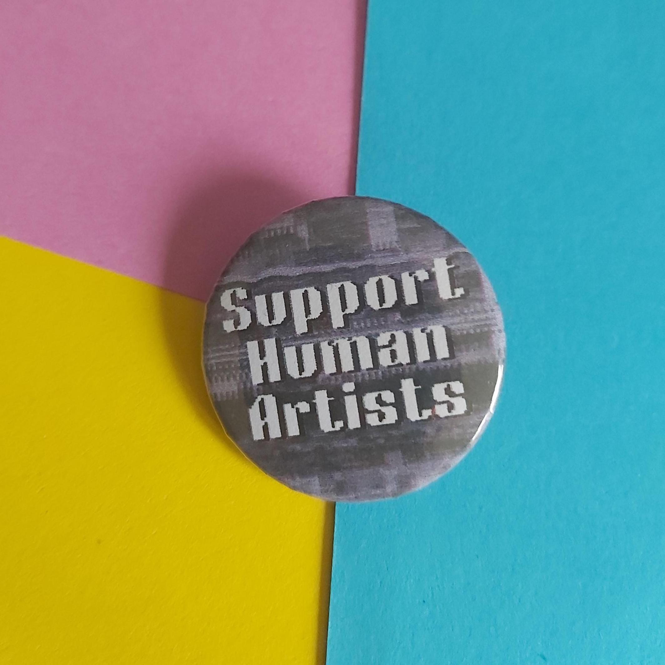 A round pinback button with the text 'Support Human Artists' in white over a gray and black abstract background, placed on a colorful background split diagonally into pink, yellow, and blue sections.