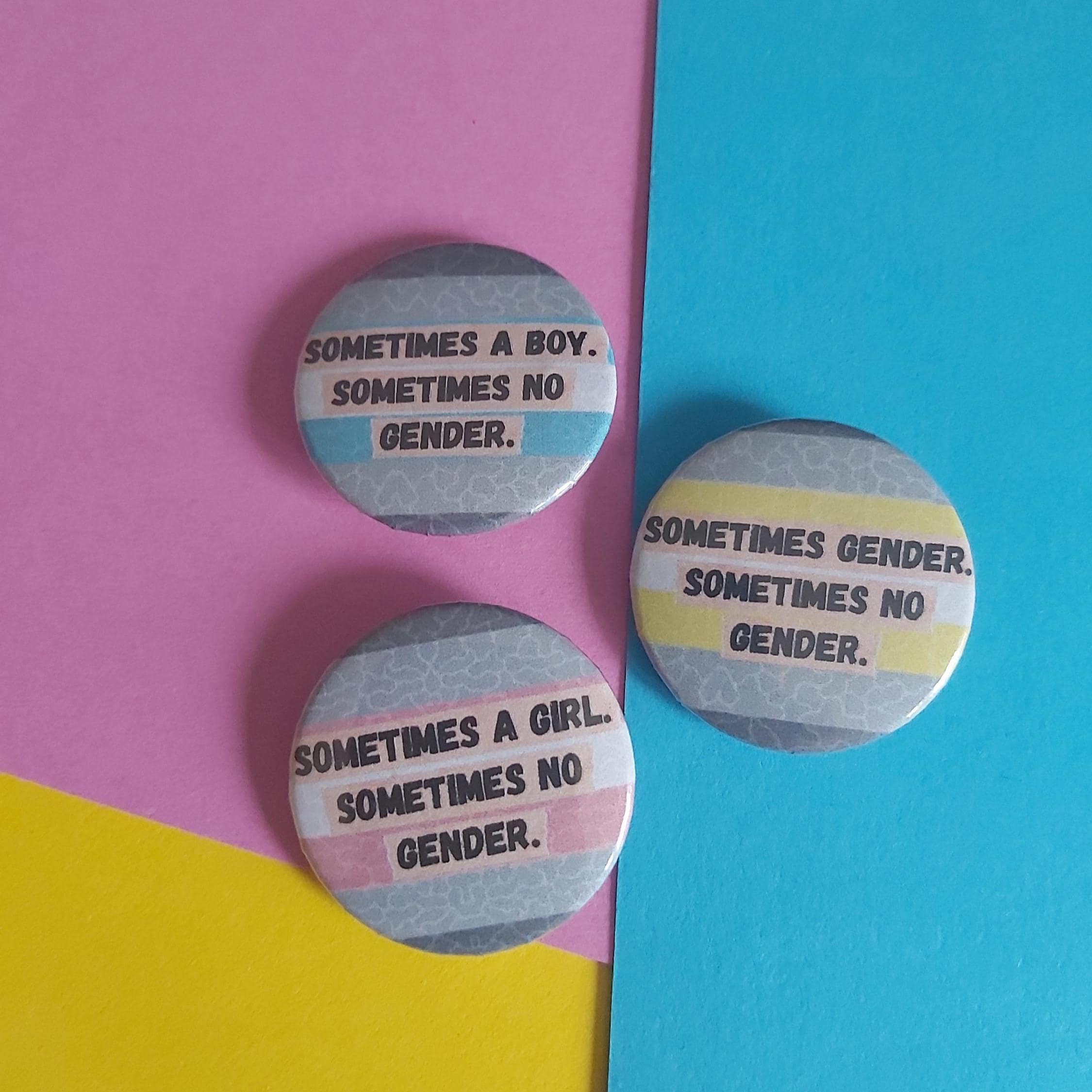 Three round pin-back buttons with gender identity text, arranged on a colourful background split diagonally between pink, blue, and yellow.