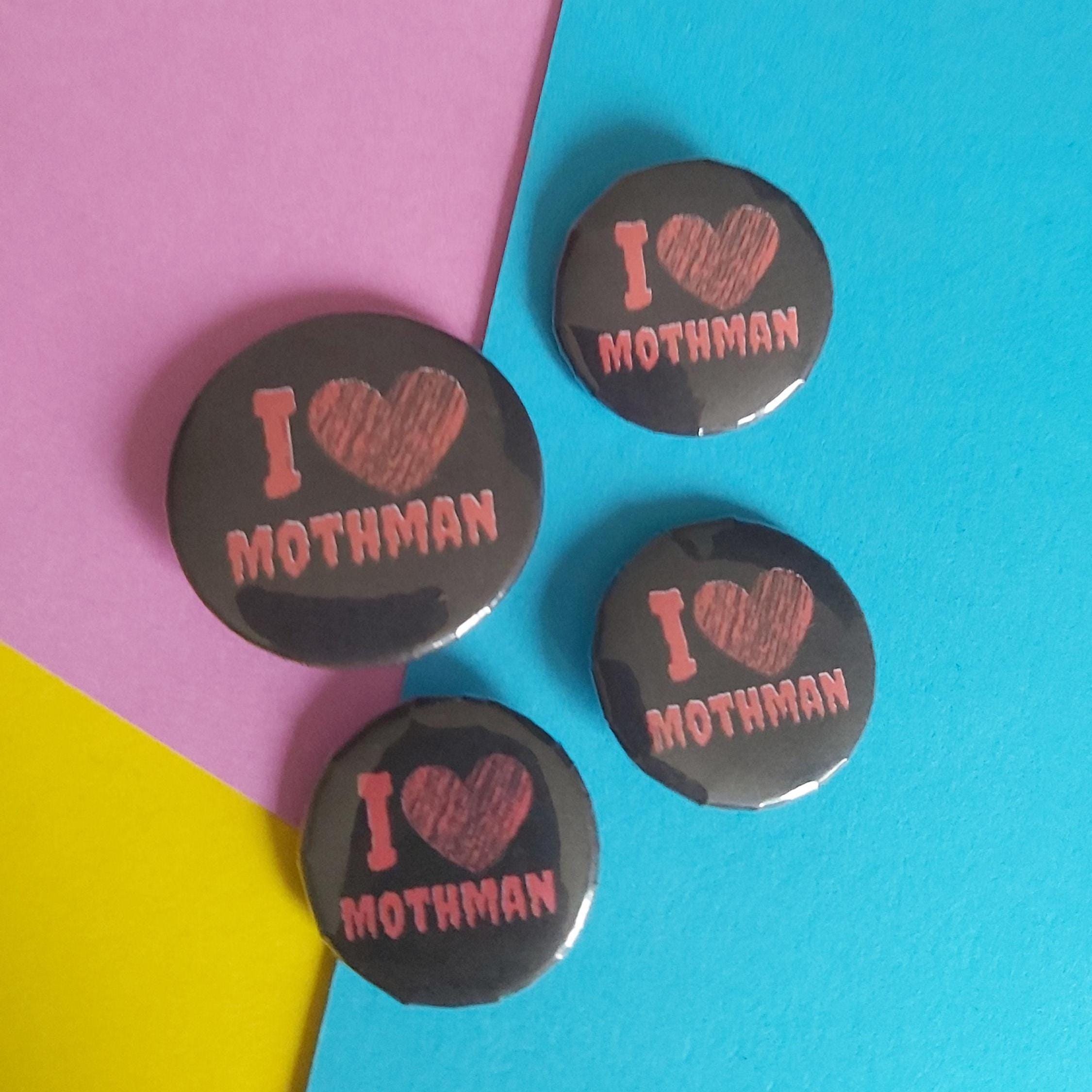 Four round pin-back buttons with a glossy black background and a pinkish-red printed design reading 'I ♥ MOTHMAN,' arranged on a colourful background split diagonally into pink, blue, and yellow sections.