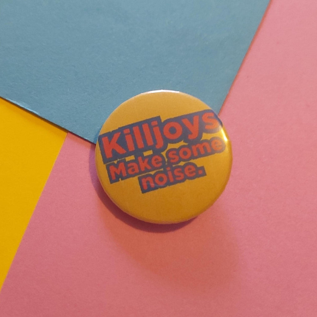 A round yellow pinback button with the text 'Killjoys Make some noise.' in red and blue, placed on a colorful background of pink, blue, and yellow paper.