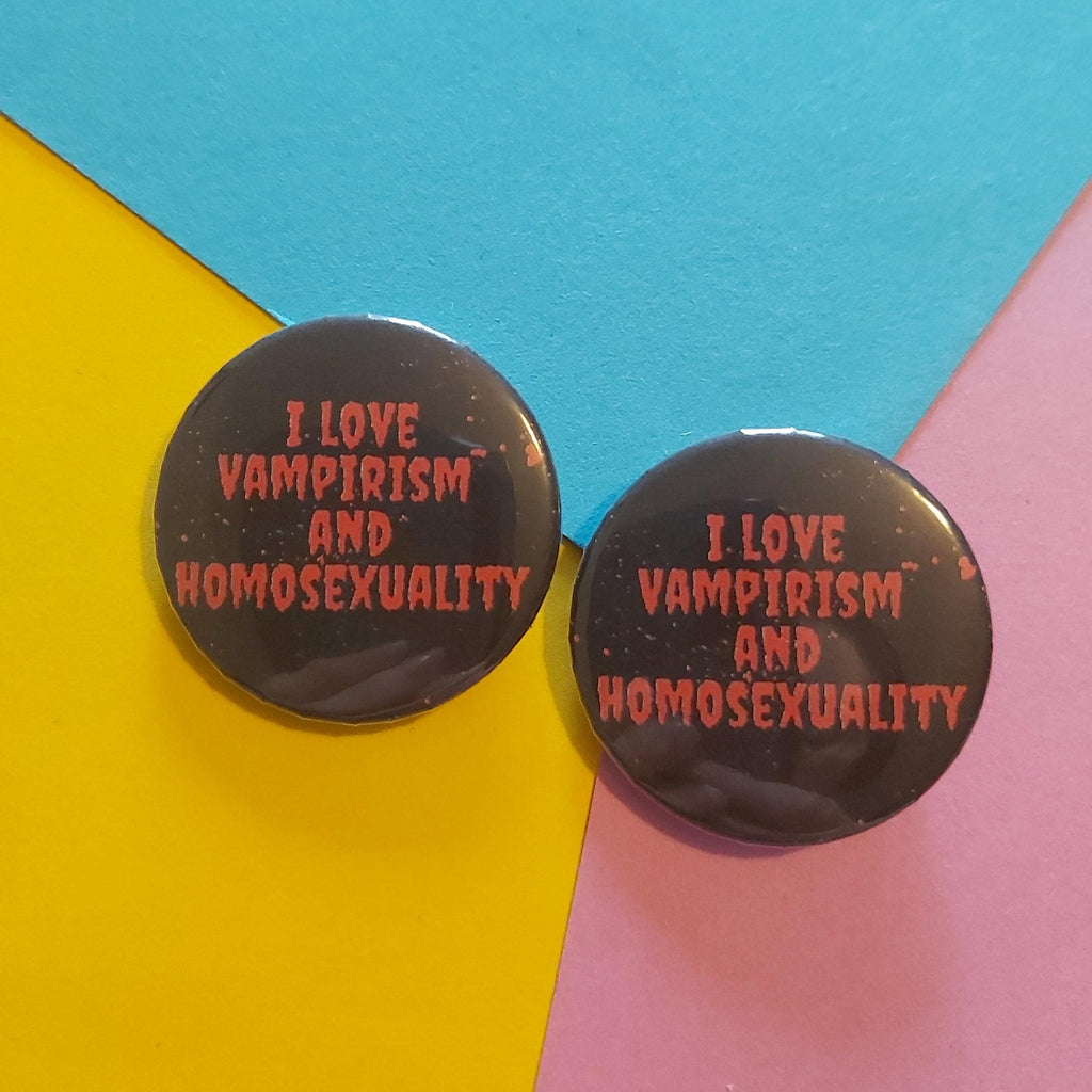 Two round black badge pins with pink text reading 'I LOVE VAMPIRISM AND HOMOSEXUALITY' displayed on a colourful background of blue, yellow, and pink paper.