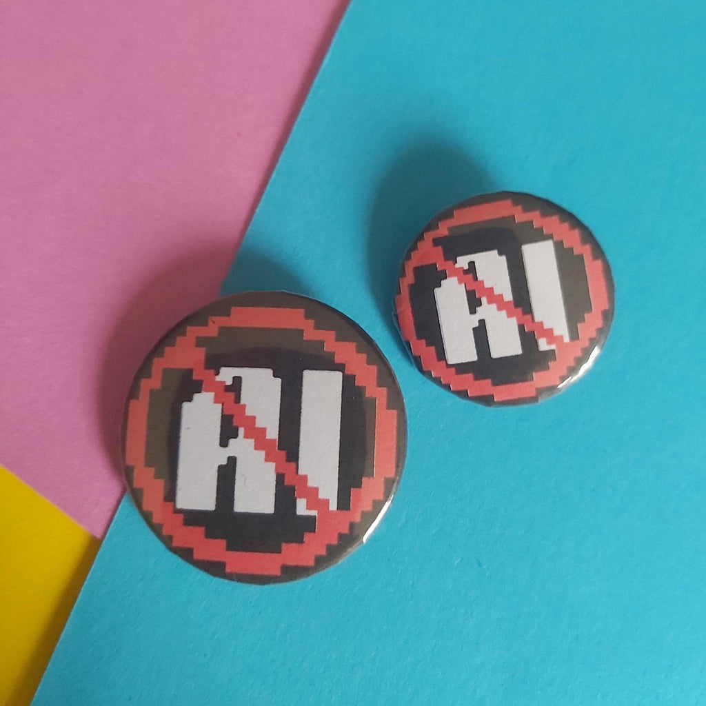 Two round pinback buttons featuring a pixel-art 'AI' text design with a red prohibition symbol over a white 'AI', displayed on a diagonally split pink, yellow, and blue background.