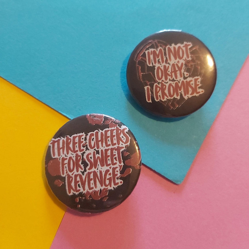 Two round pin badges on a colourful geometric background. One pin is black with the text 'THREE CHEERS FOR SWEET REVENGE' in white and red nd the other reads 'I'M NOT OKAY. I PROMISE'
