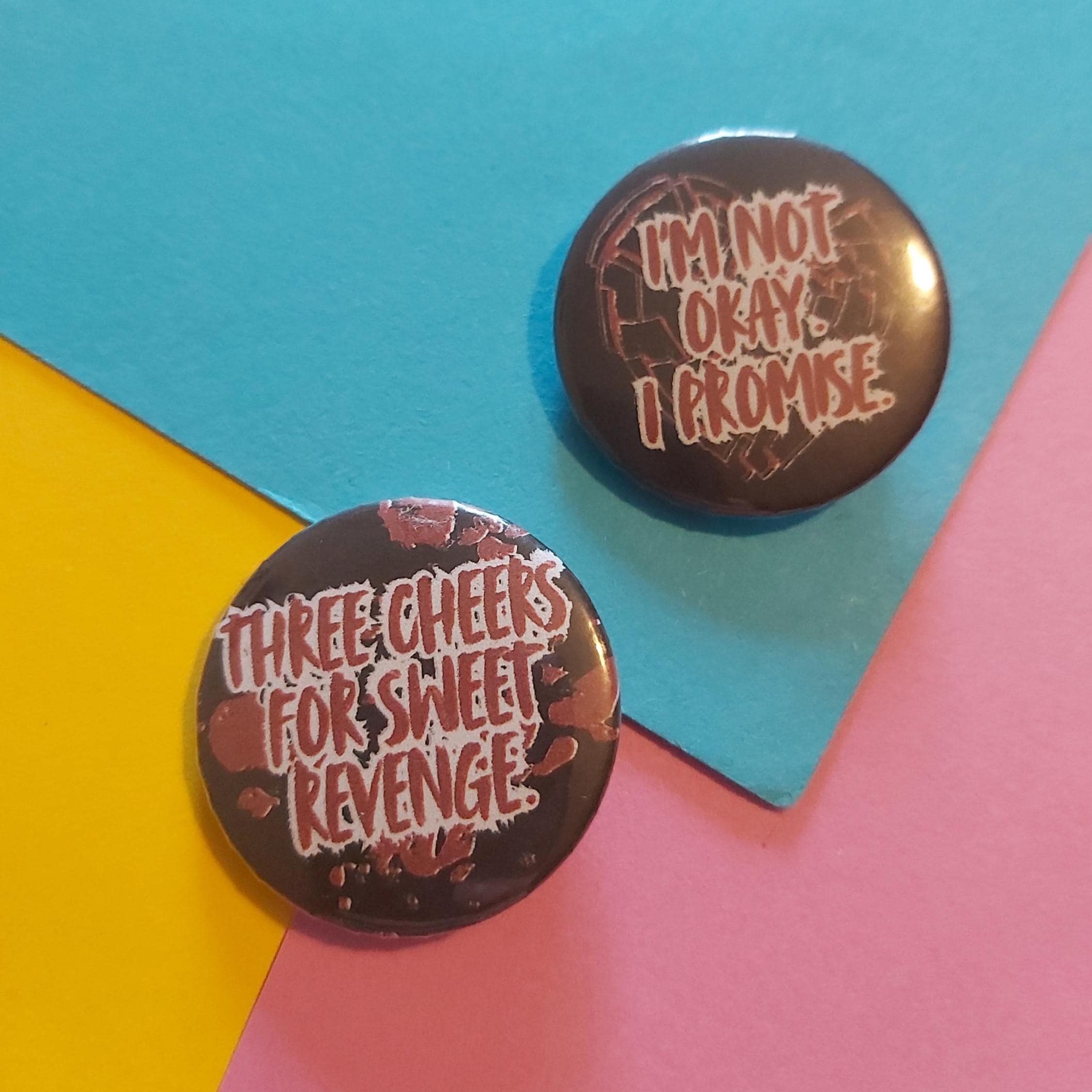 Two round pin badges on a colourful geometric background. One pin is black with the text 'THREE CHEERS FOR SWEET REVENGE' in white and red nd the other reads 'I'M NOT OKAY. I PROMISE'