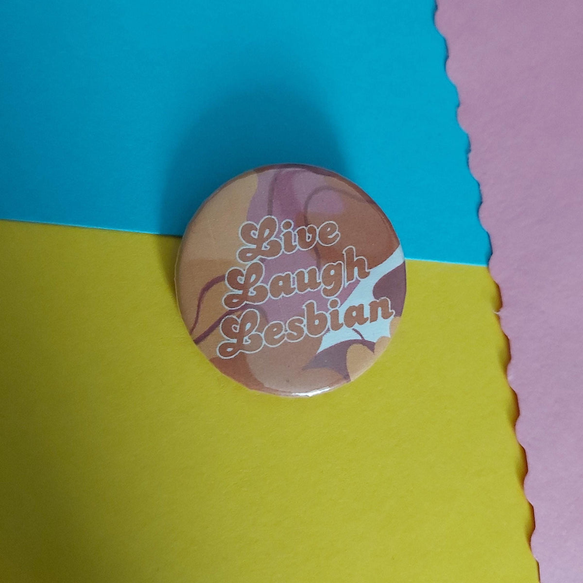 A round pinback button badge with the words 'Live Laugh Lesbian' in pink script over a marbled orange, pink, and white background, photographed on a colourful paper backdrop split between blue, yellow, and pink sections.