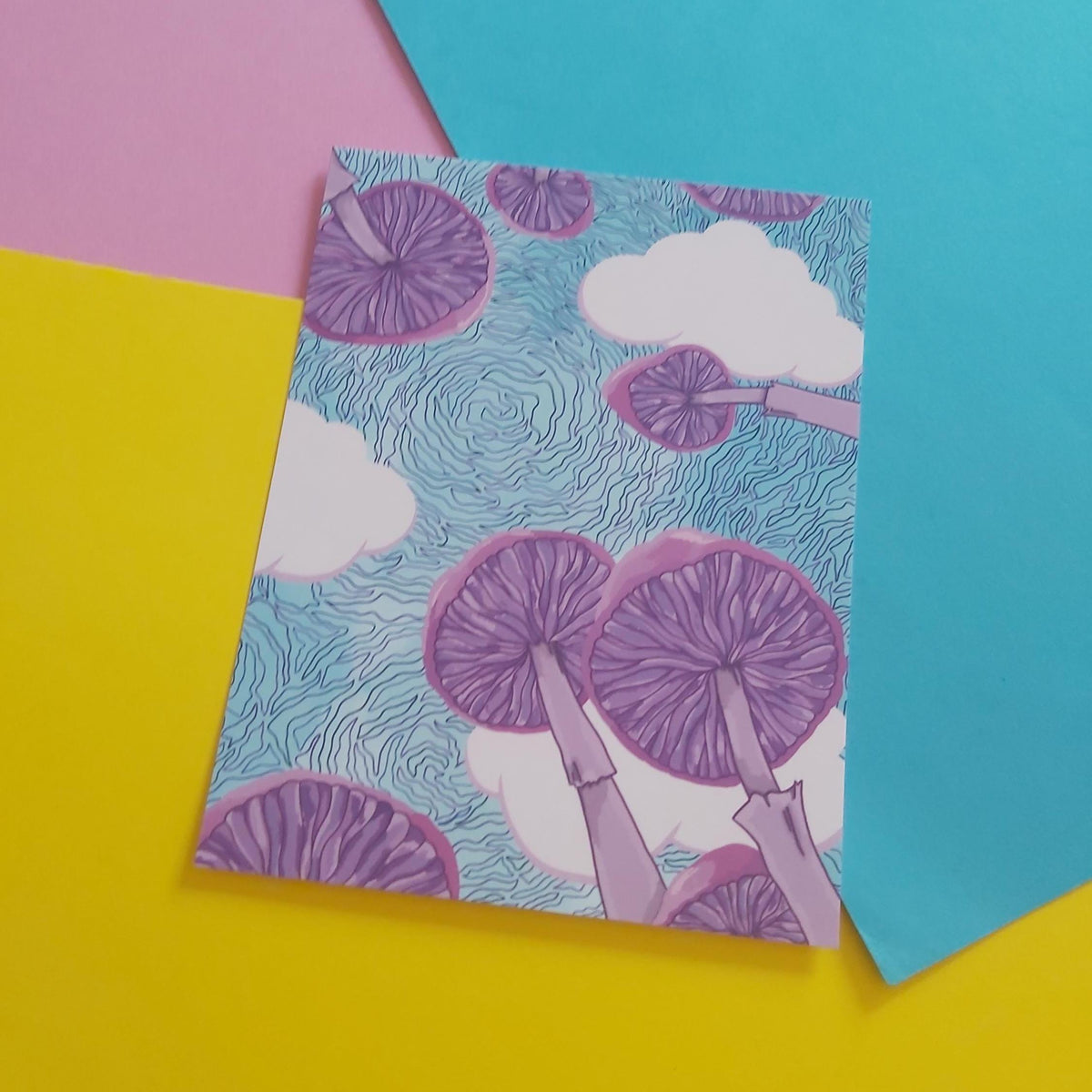 A square art print featuring a whimsical illustration of purple mushrooms reaching up toward white clouds on a light blue wavy background, styled on a colourful flat-lay surface with pink, yellow, and teal paper sections.