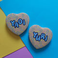 Two heart-shaped pins with a pastel heart pattern and the names 'YAOI' and 'YURI' in blue bubble letters, displayed on a colourful background of yellow, pink, and blue paper.