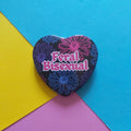 A heart-shaped pin with a dark background, pink, purple and blue floral pattern, and the text 'Feral Bisexual' in pink, placed on a diagonally split yellow, blue, and pink background.