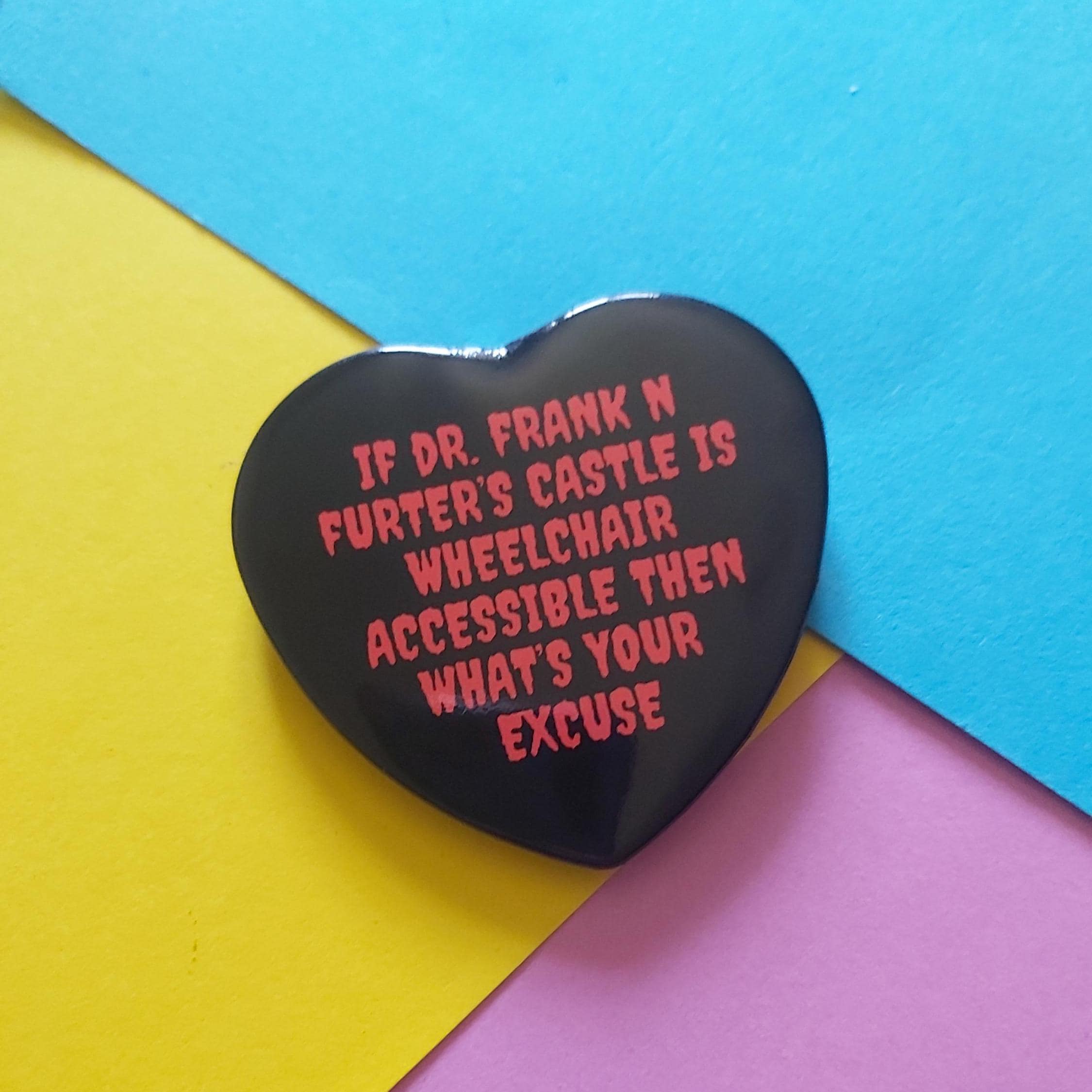 A heart-shaped black pin with red text that reads 'IF DR. FRANK N FURTER'S CASTLE IS WHEELCHAIR ACCESSIBLE THEN WHAT'S YOUR EXCUSE', displayed on a background of colored paper in blue, yellow, and pink.