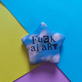 A star-shaped pin with a blue sky and white clouds pattern, featuring the text 'Fuck AI art' in black. The pin is displayed on a colourful background composed of three geometric paper shapes in blue, yellow, and purple.
