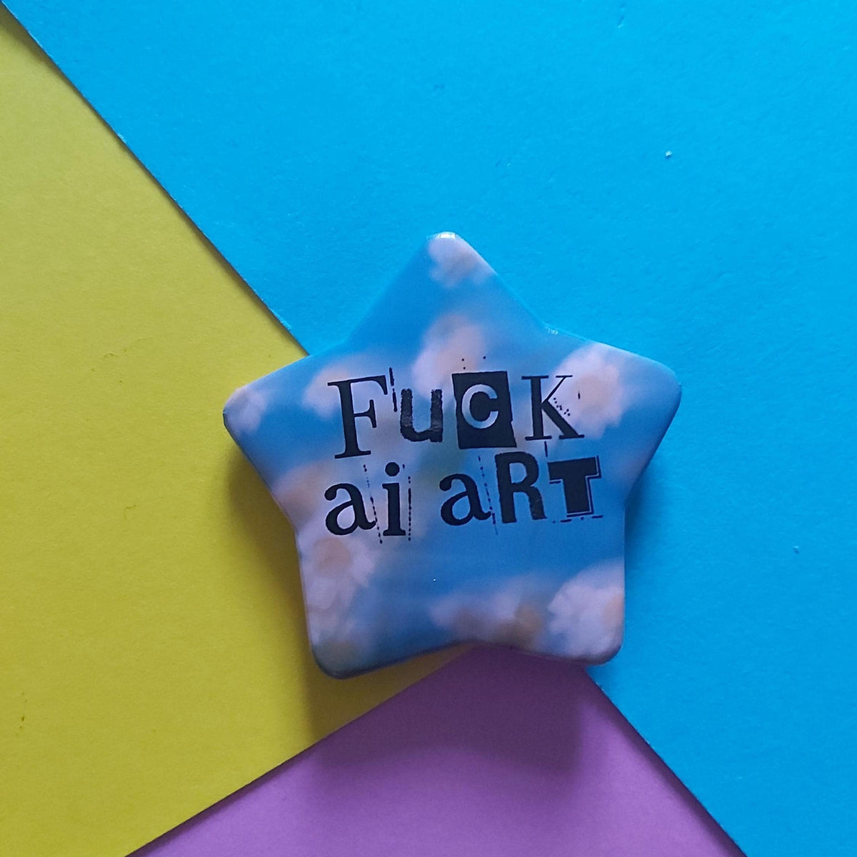 A star-shaped pin with a blue sky and white clouds pattern, featuring the text 'Fuck AI art' in black. The pin is displayed on a colourful background composed of three geometric paper shapes in blue, yellow, and purple.