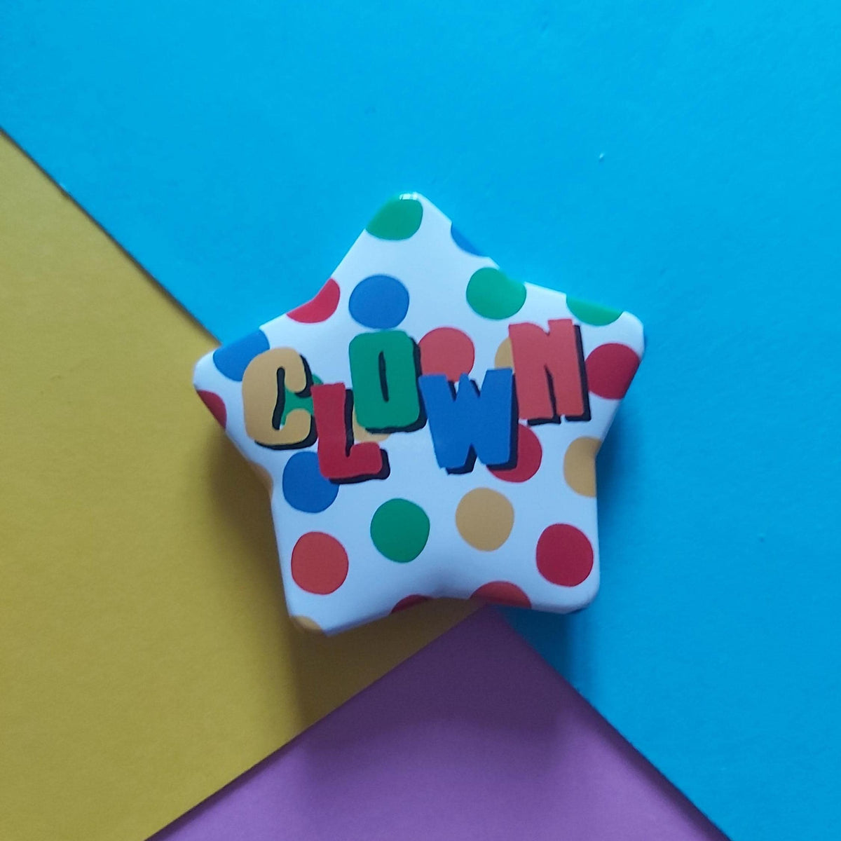 A star-shaped pin with a white background, multicoloured polka dots, and the word 'CLOWN' in colourful raised letters, displayed on a diagonally split blue, yellow, and pink background.