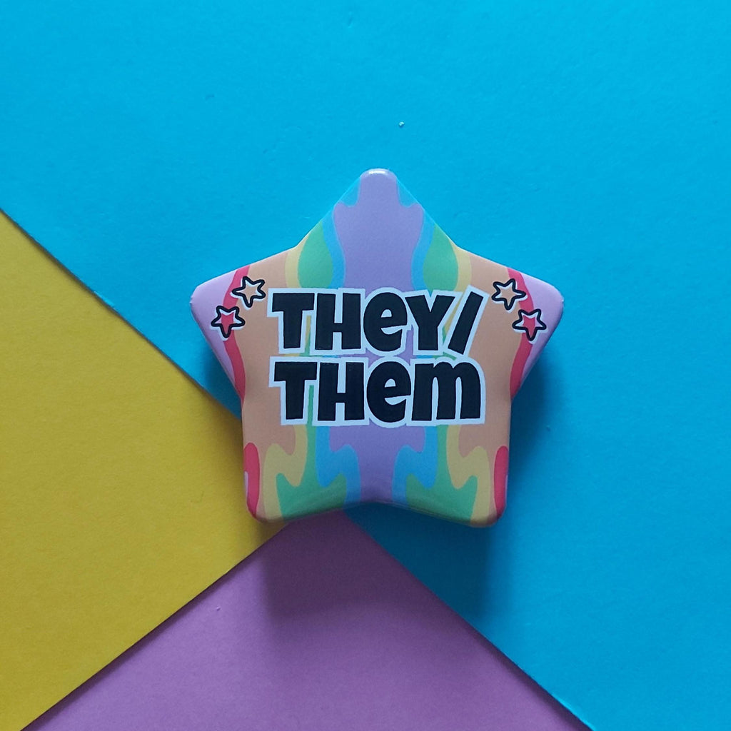 A star-shaped pin with the text 'THEY/ THEM' in black with a white outline, set against a colourful pastel tie-dye background. The pin is photographed on a background composed of three colored paper sheets: blue, yellow, and purple.