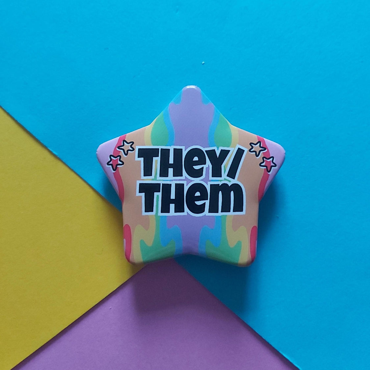 A star-shaped pin with the text 'THEY/ THEM' in black with a white outline, set against a colourful pastel tie-dye background. The pin is photographed on a background composed of three colored paper sheets: blue, yellow, and purple.