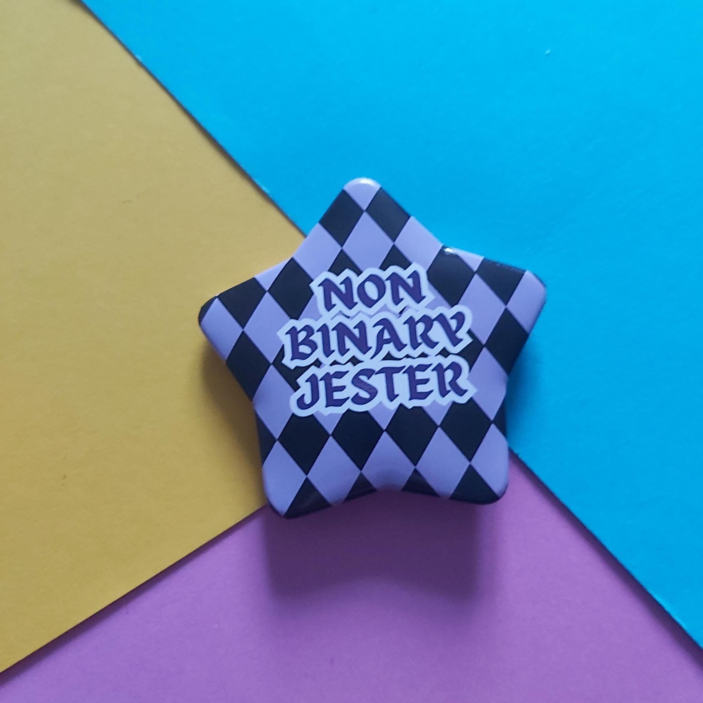 A star-shaped pin with a black and lavender checkered pattern and the text 'NON BINARY JESTER' in white and purple, placed on a diagonally split yellow, blue, and purple background.