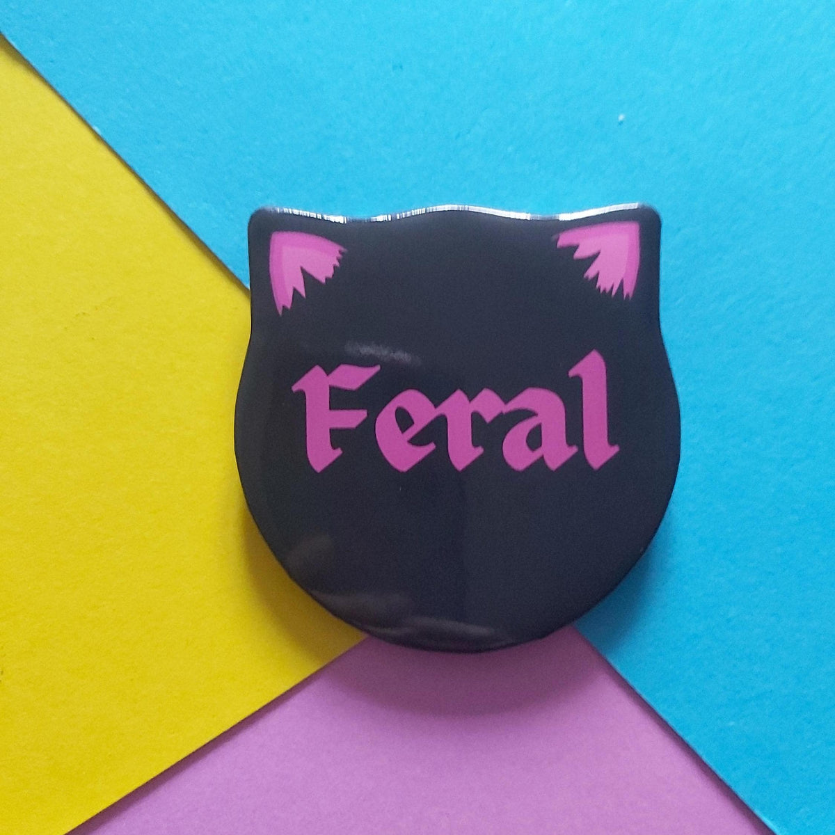 A black pin shaped like a cat head with pink ears and the word 'Feral' in pink gothic lettering, shown on a colourful background of yellow, blue, and purple paper.