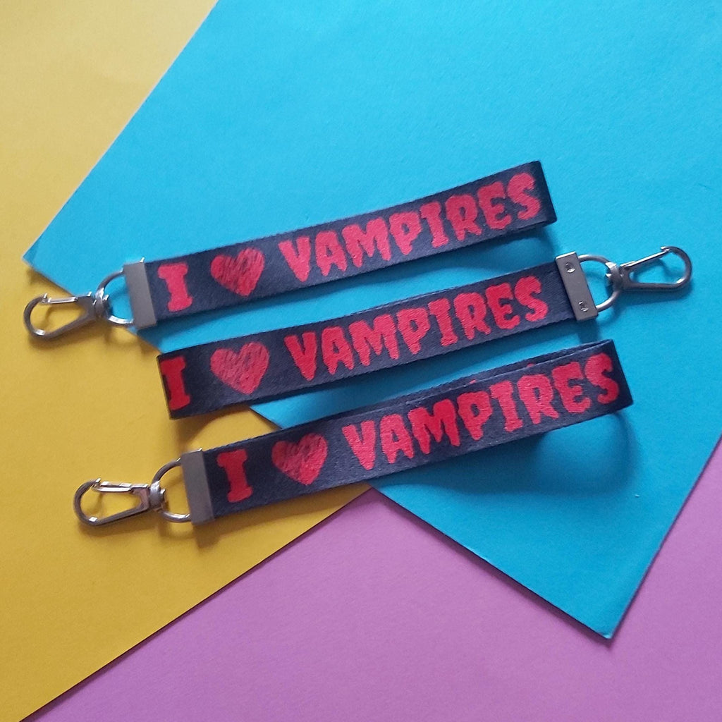 Three black keychains with silver clasps, printed with 'I ♥ VAMPIRES' in red, arranged diagonally on a colourful background of yellow, blue, and purple paper.