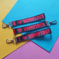 Three black keychains with silver clasps, printed with 'I ♥ VAMPIRES' in red, arranged diagonally on a colourful background of yellow, blue, and purple paper.