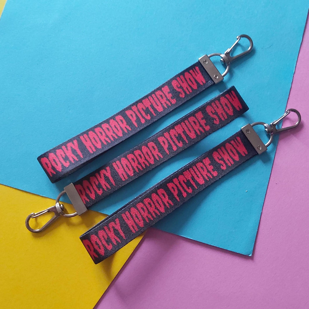 Three black fabric lanyards with silver metal clasp ends, printed with the words 'ROCKY HORROR PICTURE SHOW' in red, arranged diagonally on a colorful background of blue, yellow, and pink paper.