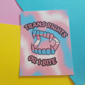 An art print featuring a stylised open mouth made of teeth with pink, blue, and white horizontal stripes, surrounded by the words 'TRANS RIGHTS OR A BITE' in pink, outlined lettering. The card is photographed on a colourful background with pink, yellow, and blue paper sections.