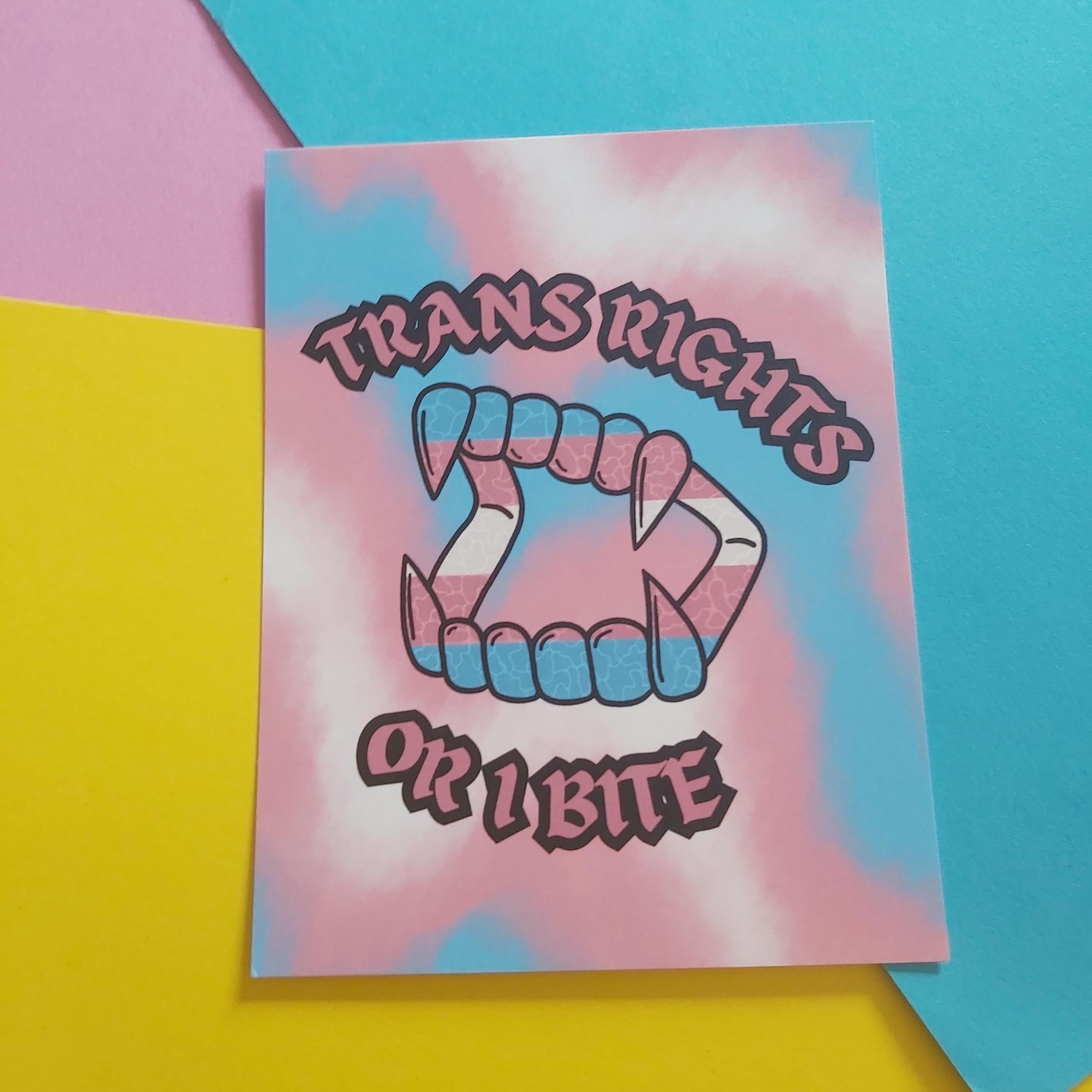 An art print featuring a stylised open mouth made of teeth with pink, blue, and white horizontal stripes, surrounded by the words 'TRANS RIGHTS OR A BITE' in pink, outlined lettering. The card is photographed on a colourful background with pink, yellow, and blue paper sections.