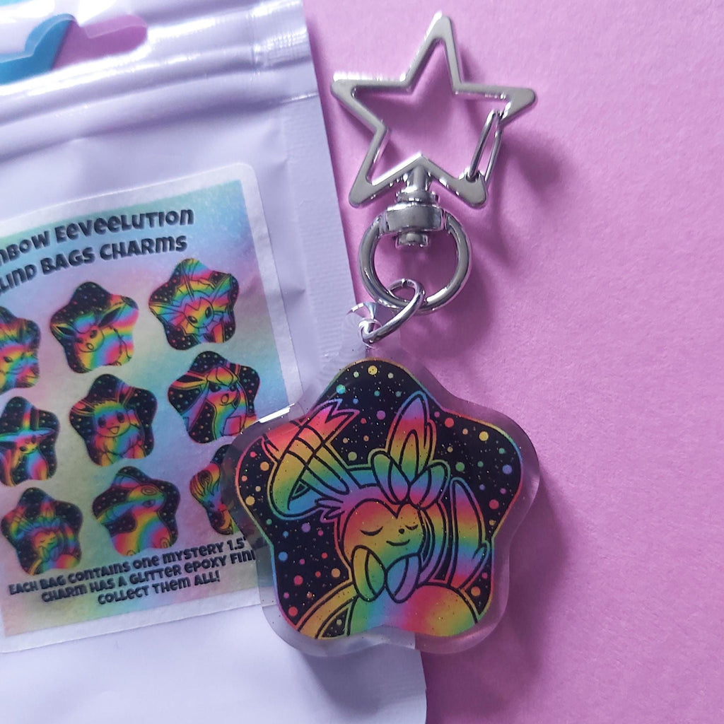 A star-shaped acrylic charm keychain with a holographic rainbow design on a black background, shown next to its packaging on a pink surface.