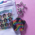 A star-shaped acrylic charm keychain with a holographic rainbow design on a black background, shown next to its packaging on a pink surface.