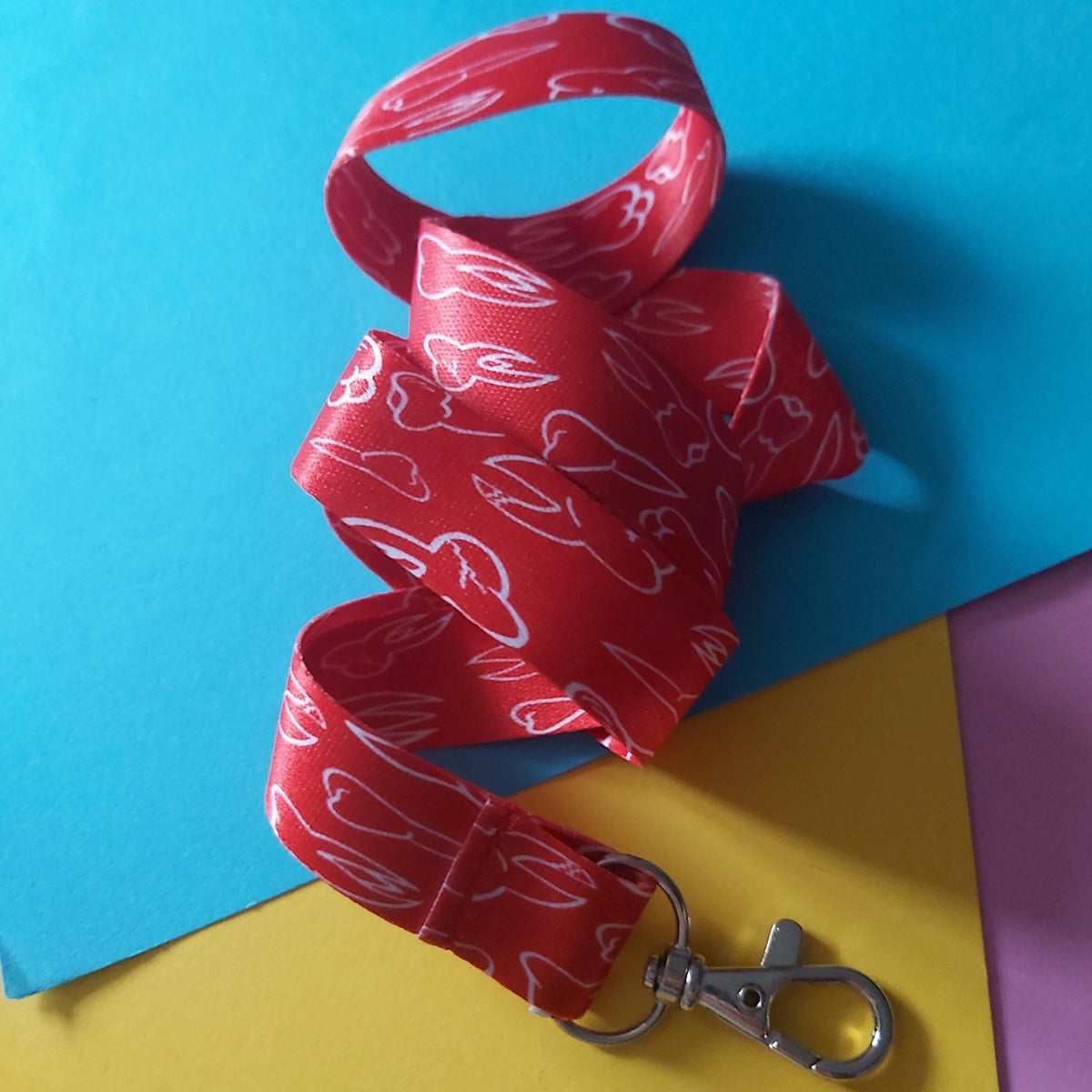 A red fabric lanyard strap with a white line-art pattern of teeth, attached to a silver metal lobster clasp, arranged on a colorful background of blue, yellow, and purple paper.