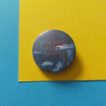 A round pin badge with the text 'NATURE ISN'T BINARY' over a dark blue and grey mushroom illustration, placed on a yellow sheet of paper over a blue background.