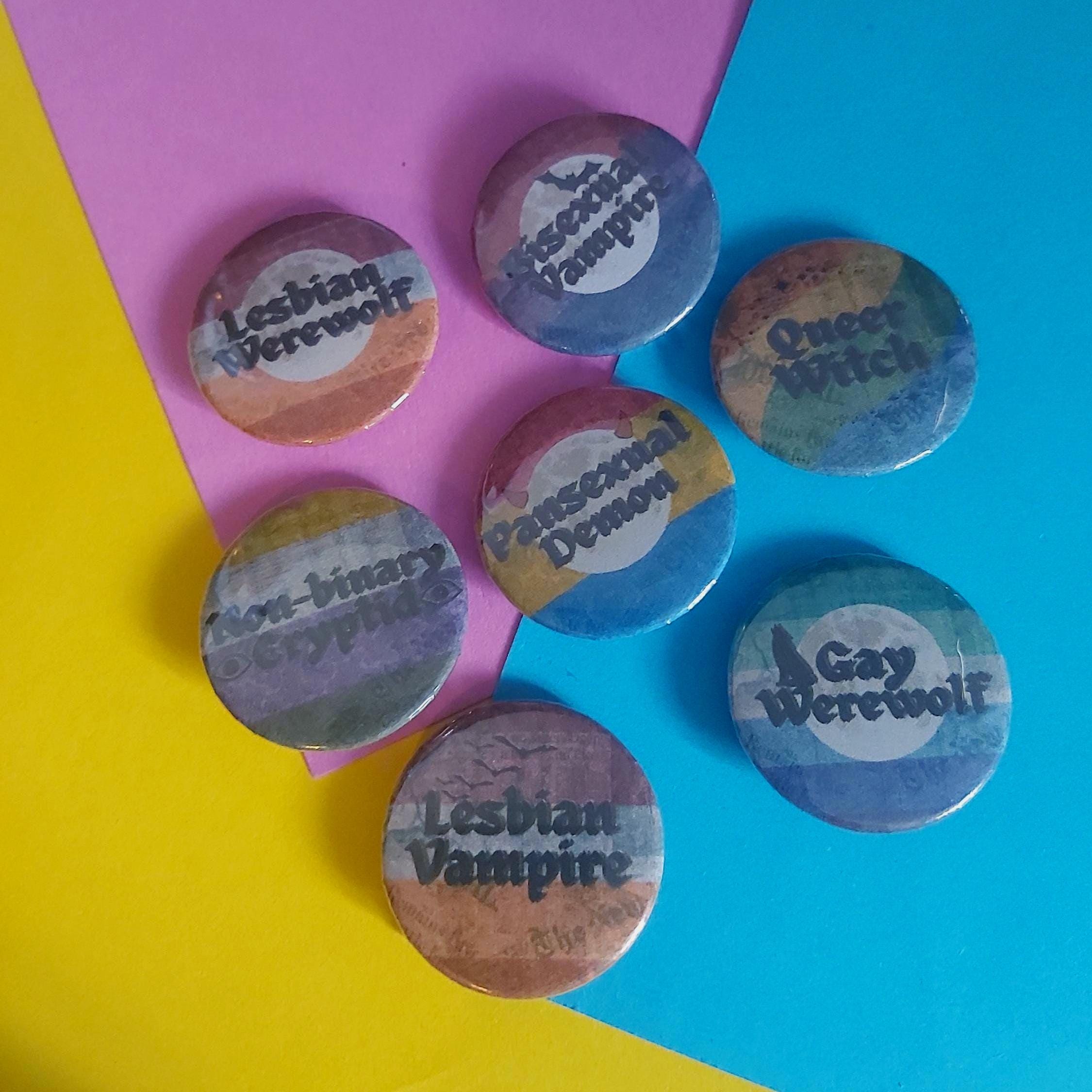 Six round pinback buttons with lesbian and queer witch-themed designs arranged on a colourful background of yellow, pink, and blue paper.
