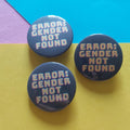 Three round pin-back buttons with the text 'ERROR: GENDER NOT FOUND' in yellow and pink on a dark background, arranged on a colourful paper backdrop with yellow, blue, and purple sections.