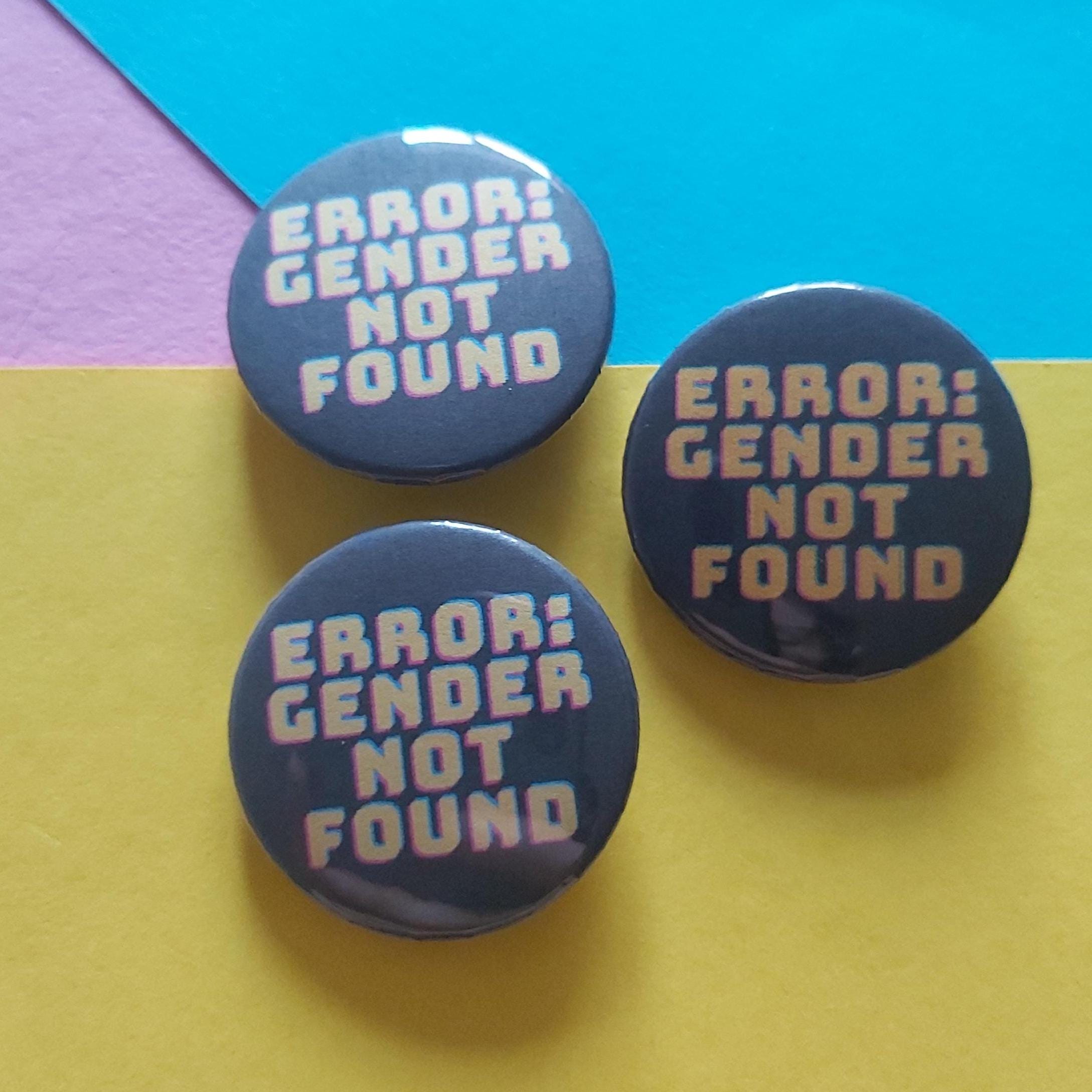 Three round pin-back buttons with the text 'ERROR: GENDER NOT FOUND' in yellow and pink on a dark background, arranged on a colourful paper backdrop with yellow, blue, and purple sections.