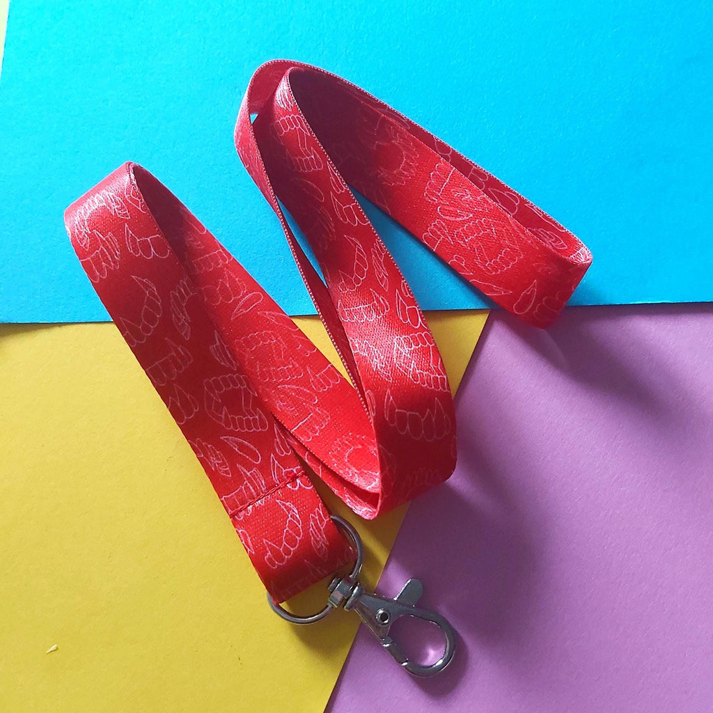 A red fabric lanyard with a repeating white line-art pattern and a silver metal lobster clasp, laid flat on a colourful background of blue, yellow, and purple paper.