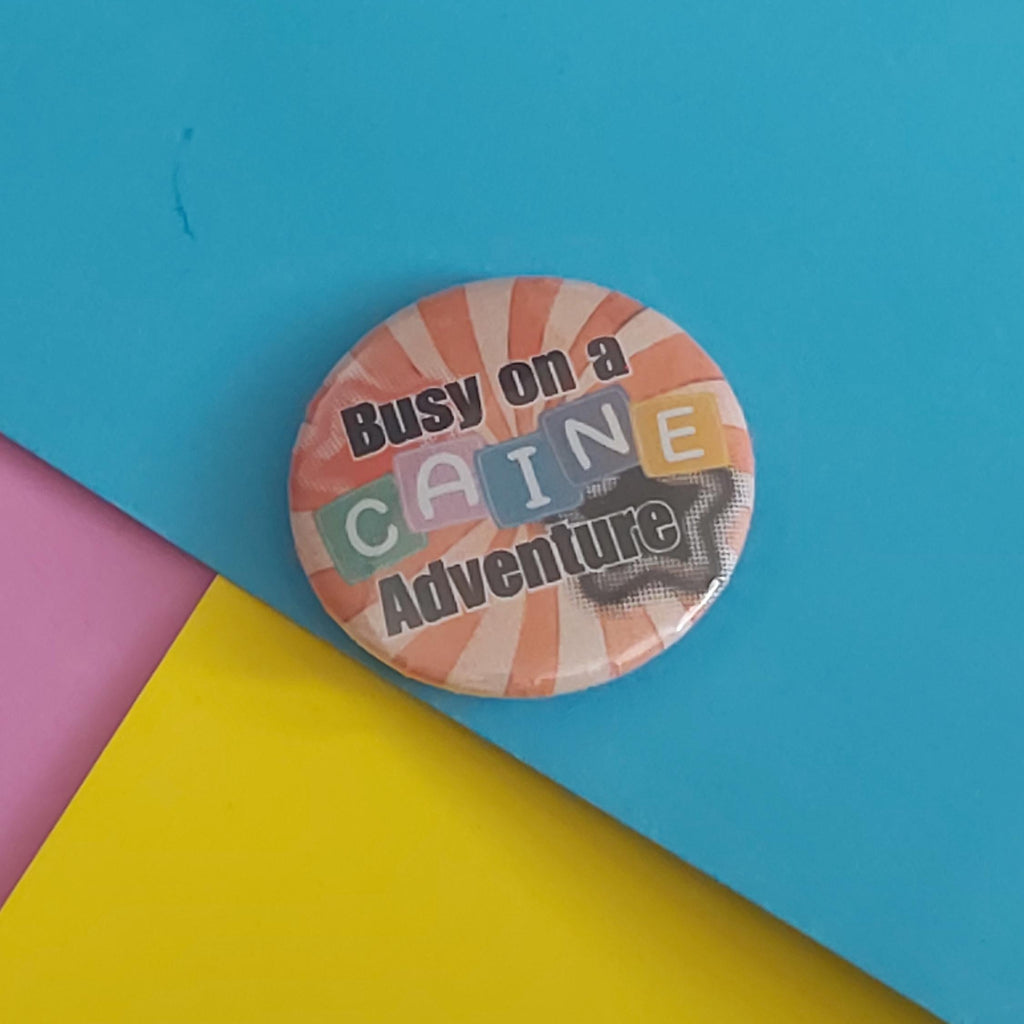 A round pin-back button badge with the text 'Busy on a CAINE Adventure' in black lettering over a retro red and white background