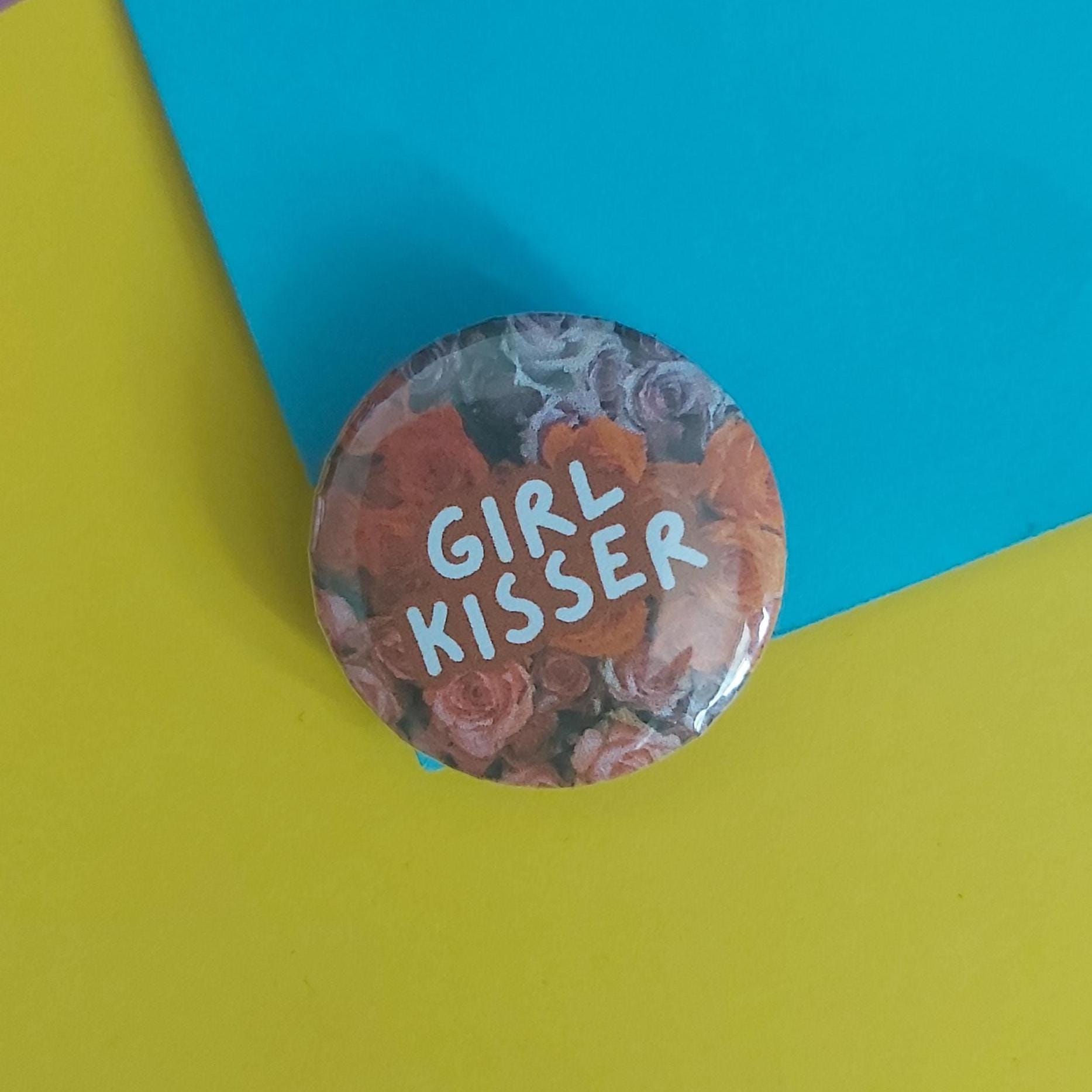 A round pinback button featuring a floral design with the text 'GIRL KISSER' in white lettering, placed on a split yellow and blue background.