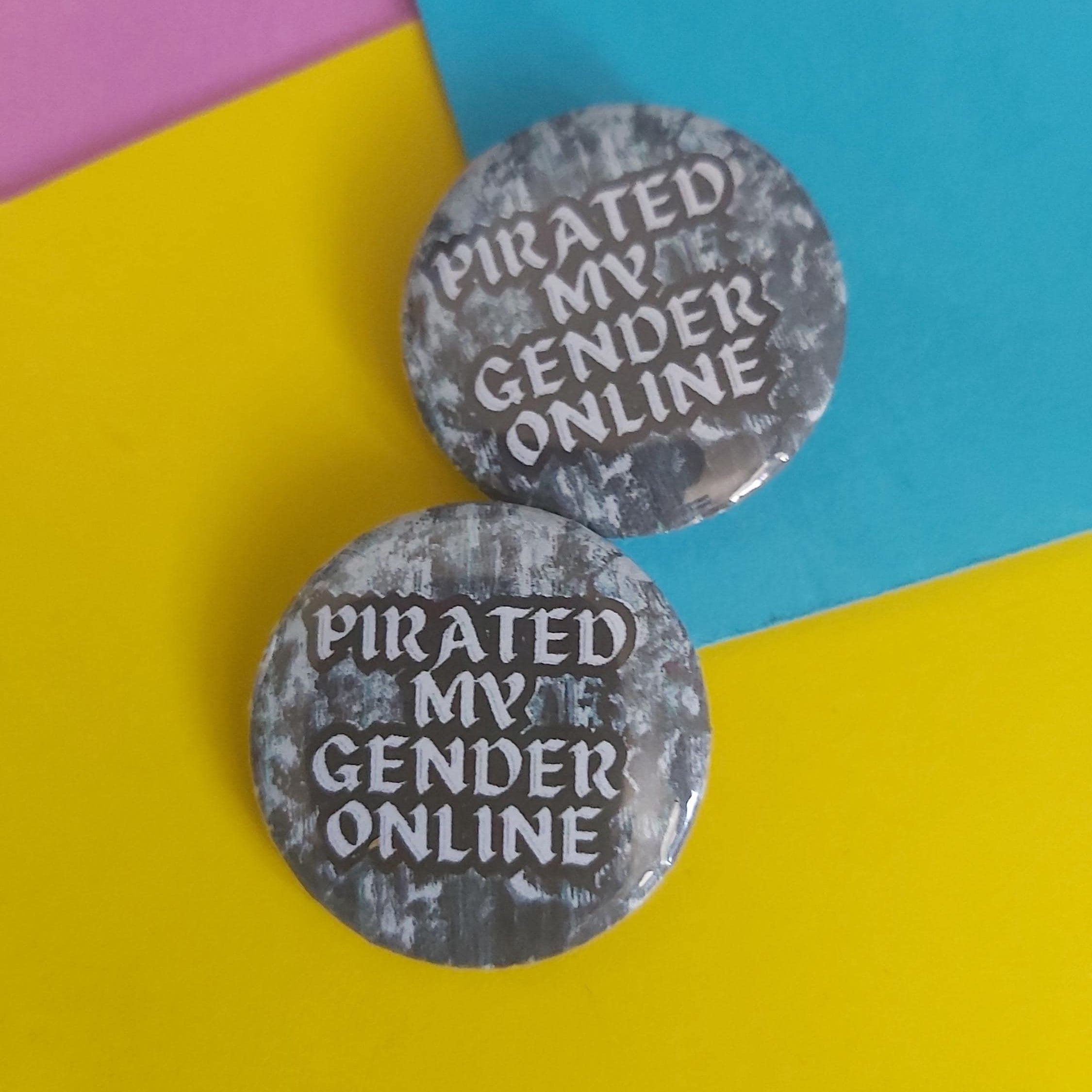 Two round grey pinback buttons with the text 'PIRATED MY GENDER ONLINE' in white, displayed on a colourful background of yellow, blue, and pink paper.