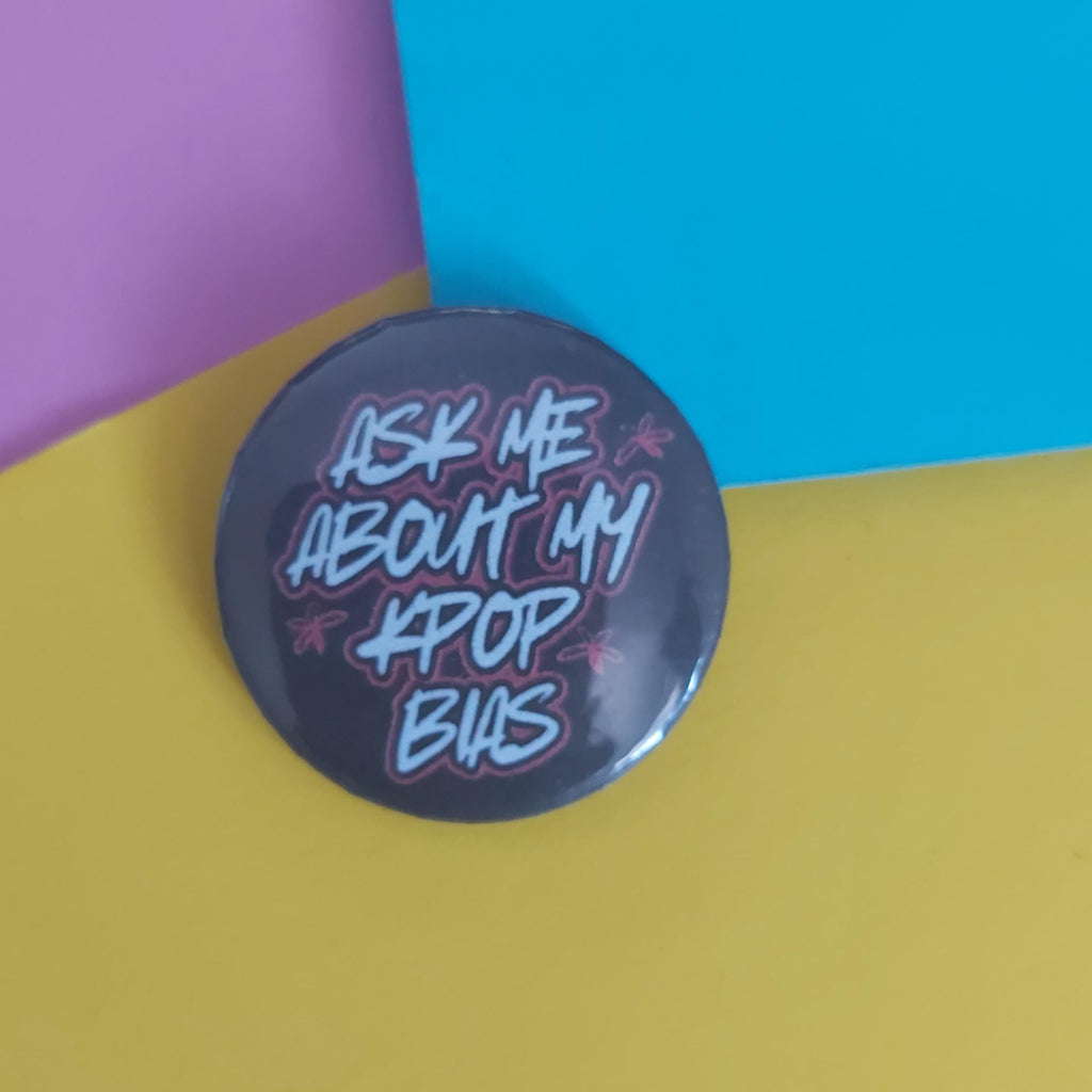 A round pinback button badge with the text 'ASK ME ABOUT MY KPOP BIAS' in stylised white and pink lettering on a dark grey background
