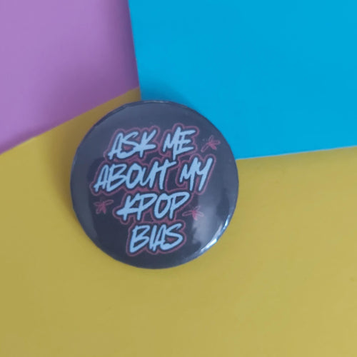 A round pinback button badge with the text 'ASK ME ABOUT MY KPOP BIAS' in stylised white and pink lettering on a dark grey background