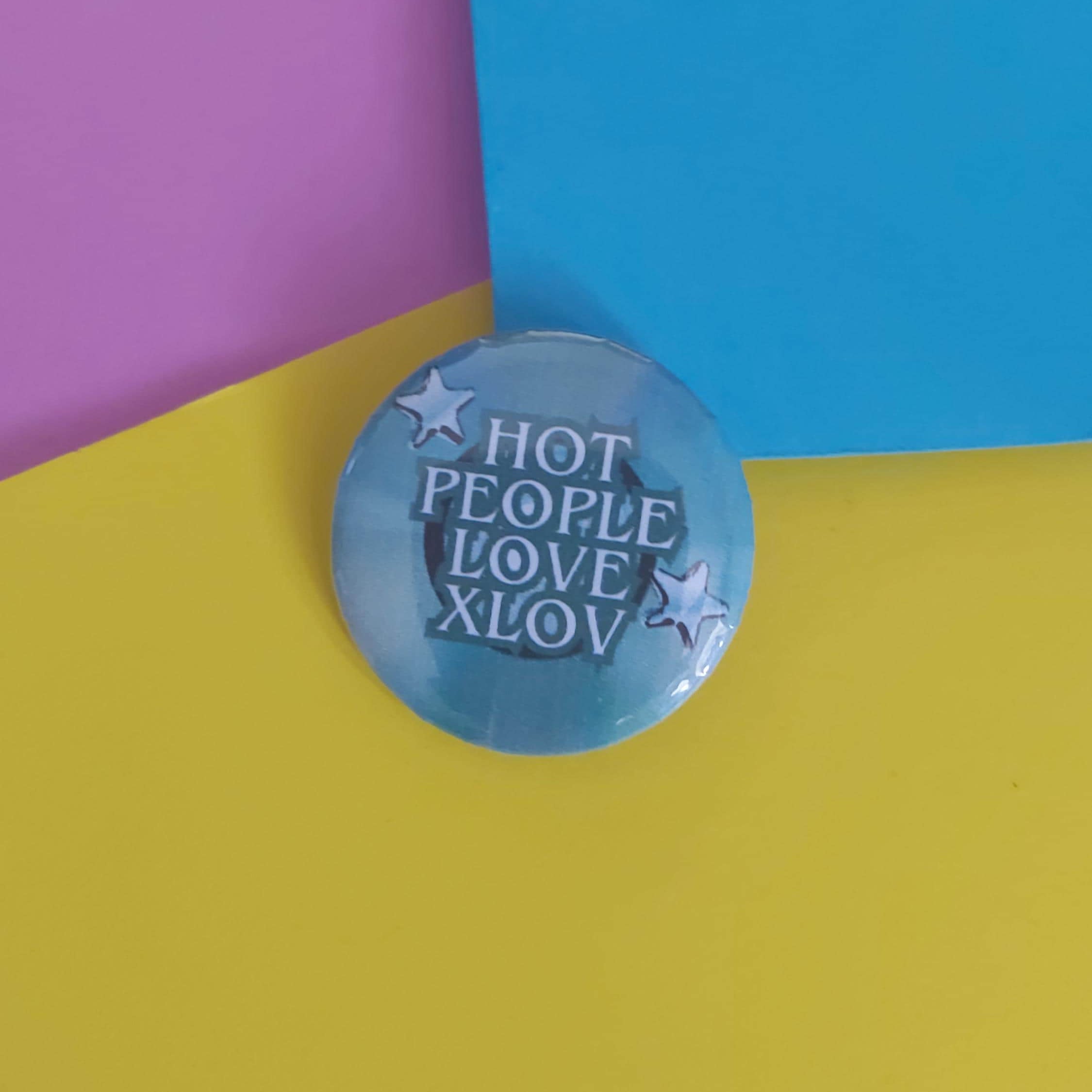 A round pinback button badge with the text 'HOT PEOPLE LOVE XLOV' in white and silver, accented with white stars, resting on a colorful background split diagonally between yellow, blue, and purple.