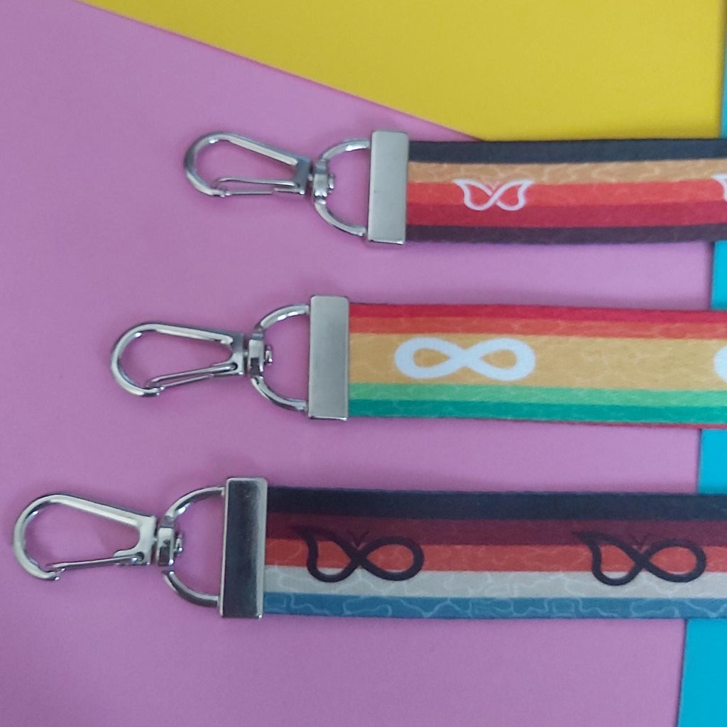 Three colourful fabric keychains with silver metal swivel clasps, arranged diagonally on a pink and yellow background.