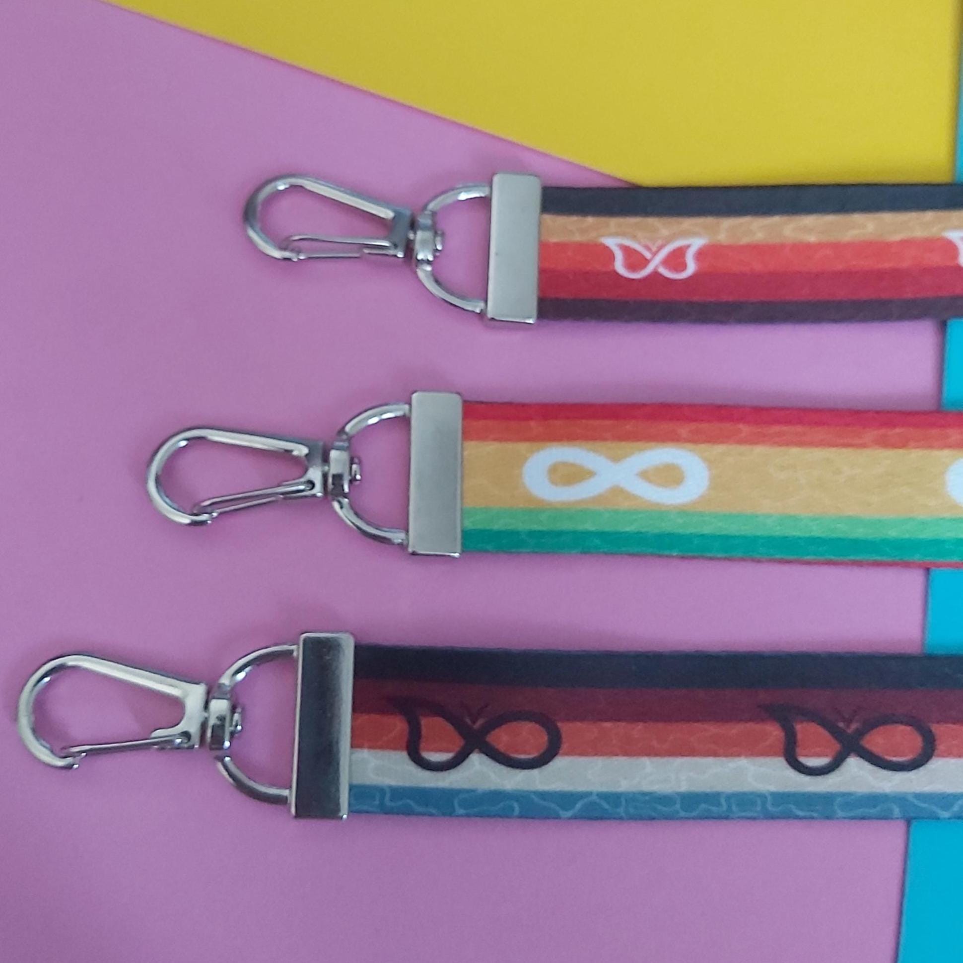 Three colourful fabric keychains with silver metal swivel clasps, arranged diagonally on a pink and yellow background.