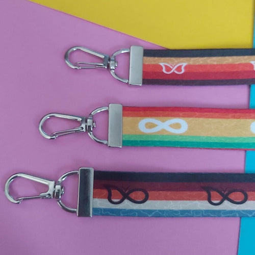 Three colourful fabric keychains with silver metal swivel clasps, arranged diagonally on a pink and yellow background.