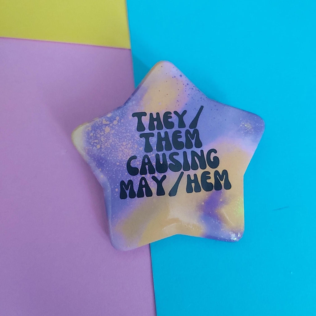 A star-shaped pin with a purple and yellow marbled design, featuring the text 'THEY / THEM CAUSING MAY/HEM' in black. The pin is displayed on a background split diagonally between pastel pink and bright cyan blue.