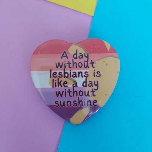 A heart-shaped enamel pin with a rainbow-striped background and the text 'A day without lesbians is like a day without sunshine' in black