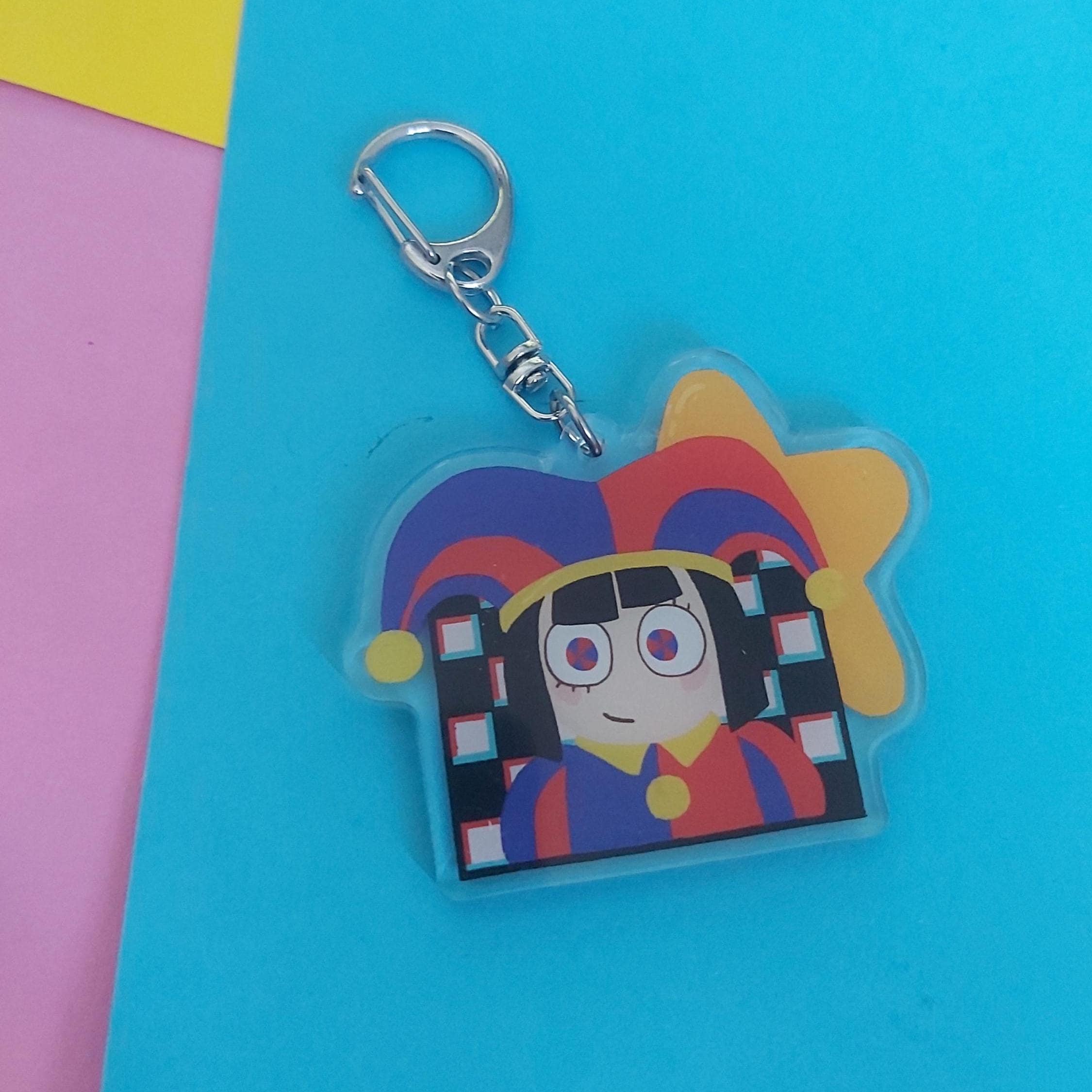 A colourful acrylic keychain featuring a cartoon character with a jester hat, attached to a silver metal clasp, displayed on a bright blue background with pink and yellow corner accents.