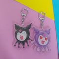 A reversible, cute cartoon keychain of Kuromi with a silver lobster clasp, one black and one purple, displayed on a pink background with yellow and blue corner accents.
