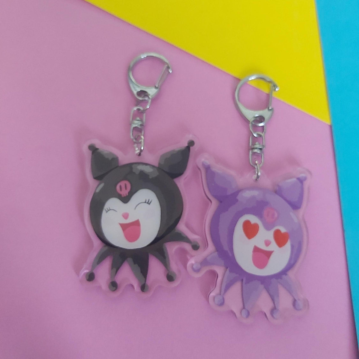 A reversible, cute cartoon keychain of Kuromi with a silver lobster clasp, one black and one purple, displayed on a pink background with yellow and blue corner accents.