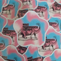 A pile of die-cut stickers featuring a pink and blue tie-dye background, a black line drawing of a dinosaur, and the text 'MAKE TRANSPHOBIA EXTINCT'.